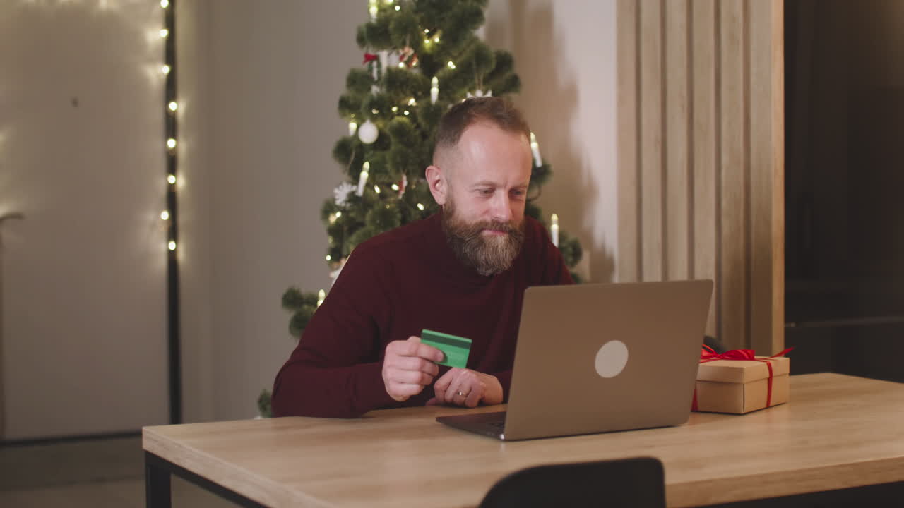 hombre pelirrojo comprando en línea con una tarjeta de crédito usando una computadora portátil sentada en una mesa cerca de un regalo en una habitación decorada con un árbol de navidad 1