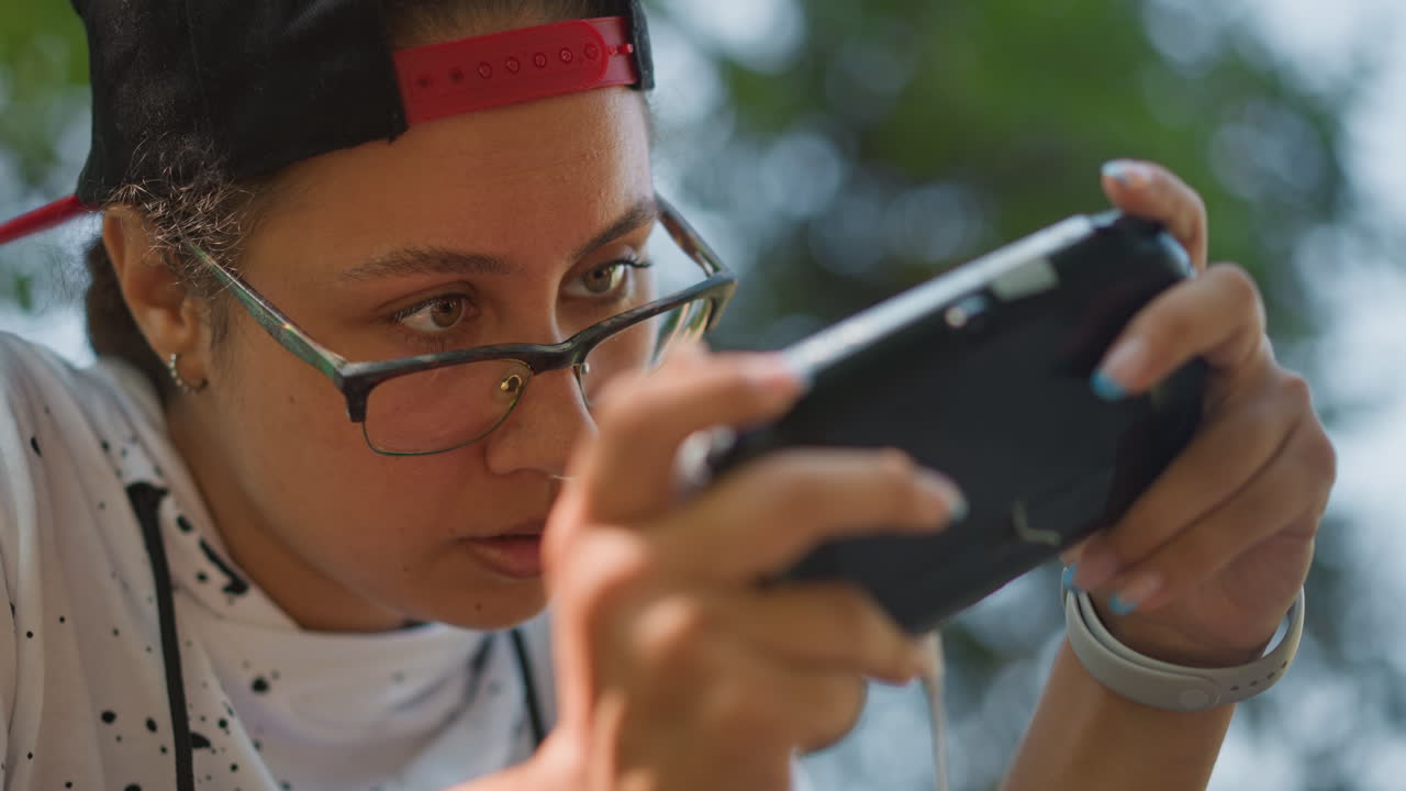 primer plano intenso de una mujer jugando al aire libre con una consola portátil en las manos, gafas y gorra roja hacia atrás, bokeh de árbol iluminado por el sol, expresión concentrada, dedos en los botones, reloj de pulsera visible, camiseta moteada