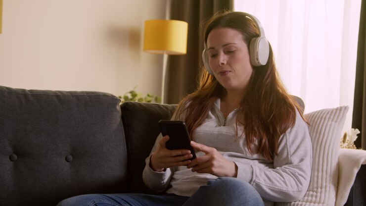 Kvinde med trådløse hovedtelefoner sidder i sofaen derhjemme og streamer musik fra mobiltelefon 2