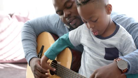 happy african american father and son sitting on sofa and playing guitar, in slow motion