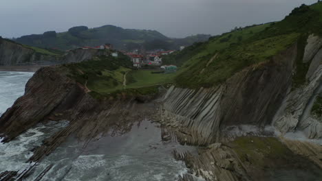 Panoramic-aerial,-itzurun-beach-cliff-side-flysch-stratigraphy-and-water-reflection-cloudy-sky