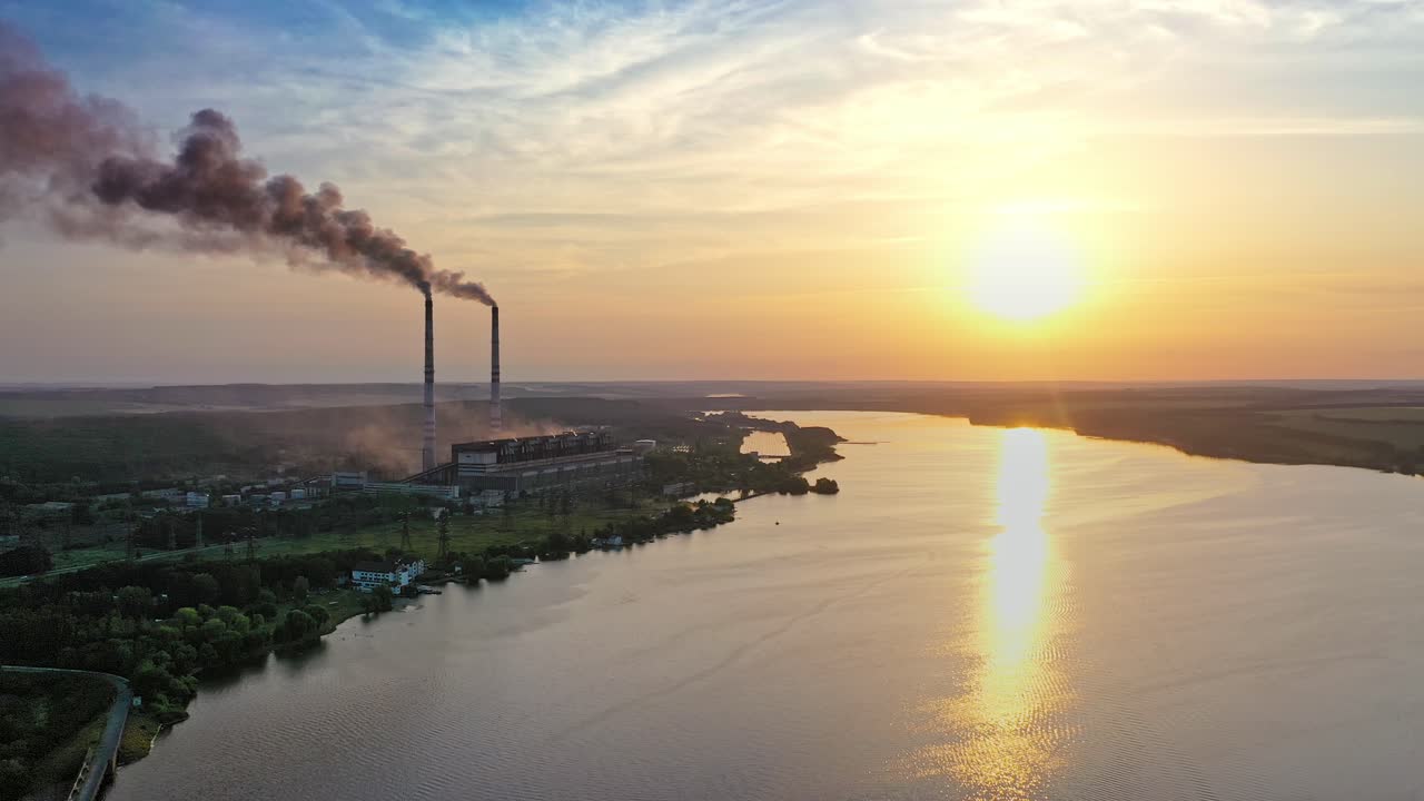 Dark smoke from industrial pipes near the water surface at sunset. Natural evening landscape of beautiful river and a factory. Pollution of the environment.