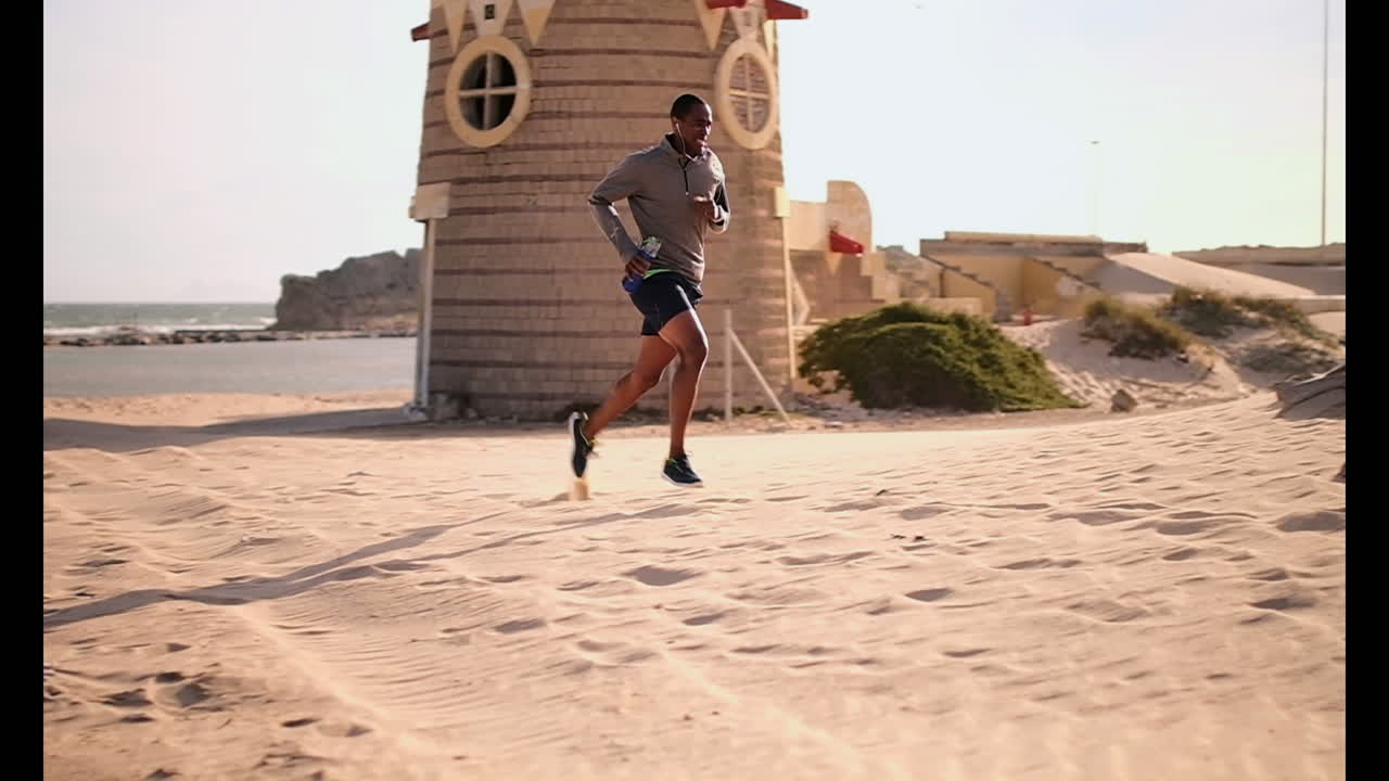 아프리카계 미국인 남성 달리기 선수가  ⁇ 빛 아래 해변에서 조 ⁇ 을 하고 있습니다.