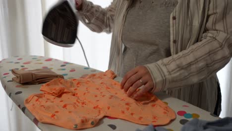 pregnant woman ironing baby clothes while touching her belly at home