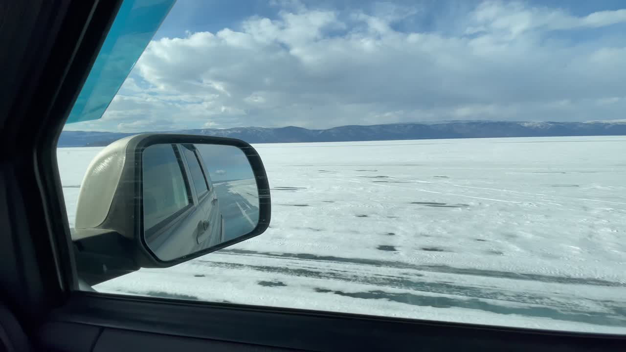 Filmato dall'interno di un'auto che guida sulla strada di ghiaccio congelata attraverso il Lago Baikal, con un'ampia superficie innevata, montagne distanti e nuvole imponenti, Russia, Siberia.