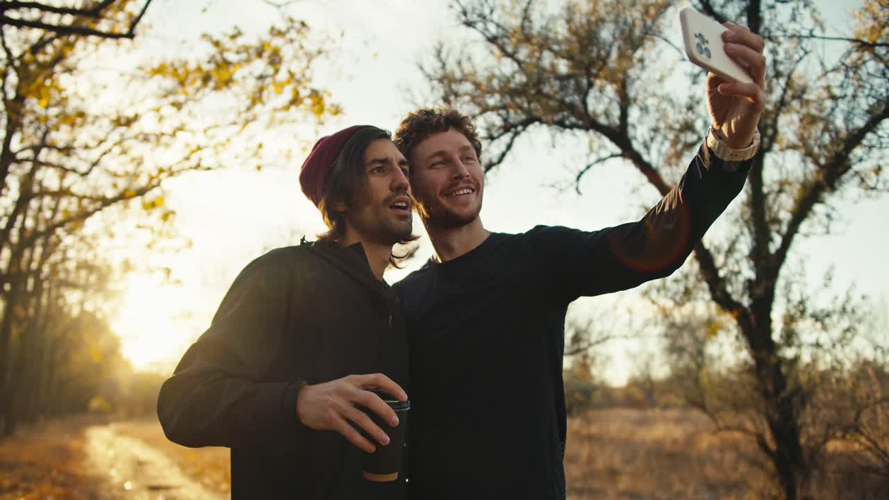 hombre feliz con el cabello rizado en un uniforme deportivo negro toma una selfie con su amigo masculino en una gorra roja con una taza de cartón de café cerca de un bosque de otoño con follaje marrón por la mañana al amanecer