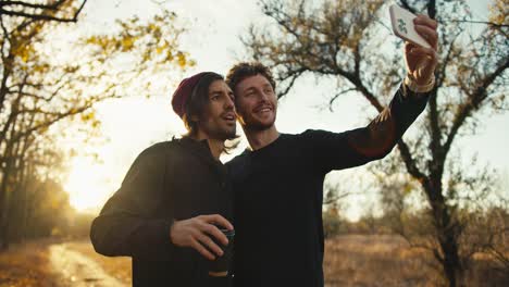 Happy-man-with-curly-hair-in-a-black-sports-uniform-takes-a-selfie-with-his-male-friend-in-a-red-cap-with-a-cardboard-cup-of-coffee-near-an-autumn-forest-with-brown-foliage-in-the-morning-at-sunrise