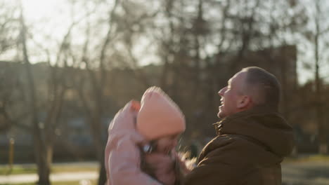 a little girl in a pink cap and jacket being joyfully raised up in the air by a man wearing a brown jacket, as they both laugh together. the scene captures a playful and happy moment in a sunlit park