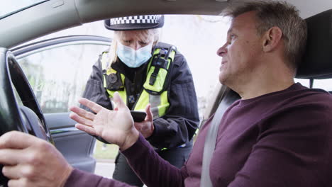 Male-Driver-Being-Stopped-By-Female-Traffic-Police-Officer-Wearing-Mask-Taking-Notes-On-Mobile-Phone