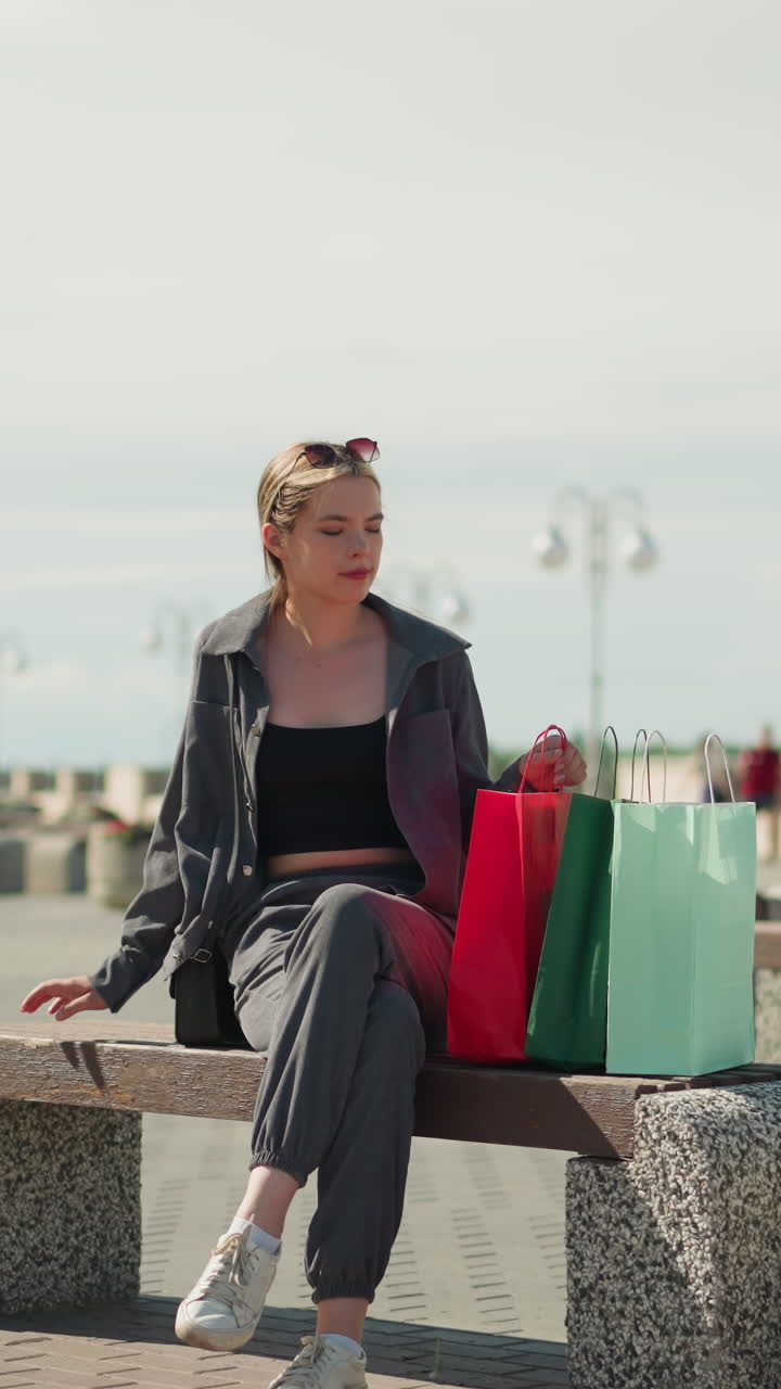 la señora se sienta en un banco al aire libre, deja caer sus bolsas de compras, y ajusta la bolsa de compras roja, mira hacia la derecha con una expresión relajada, rodeada de bancos de parque, flores y farolas