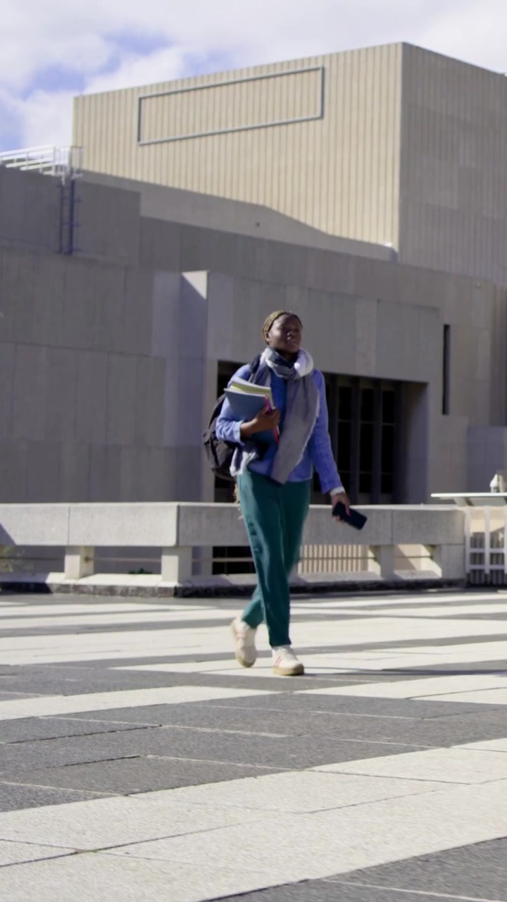 Vertical video: Entering left, student with bag and notes crossing plaza, checking phone for class