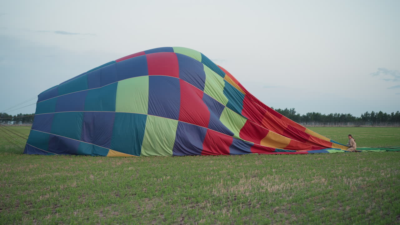 woman and boy folding vibrant multicolor hot air balloon fabric on green field at dusk while securing envelope mouth with rope during careful landing pack up under soft pastel sky