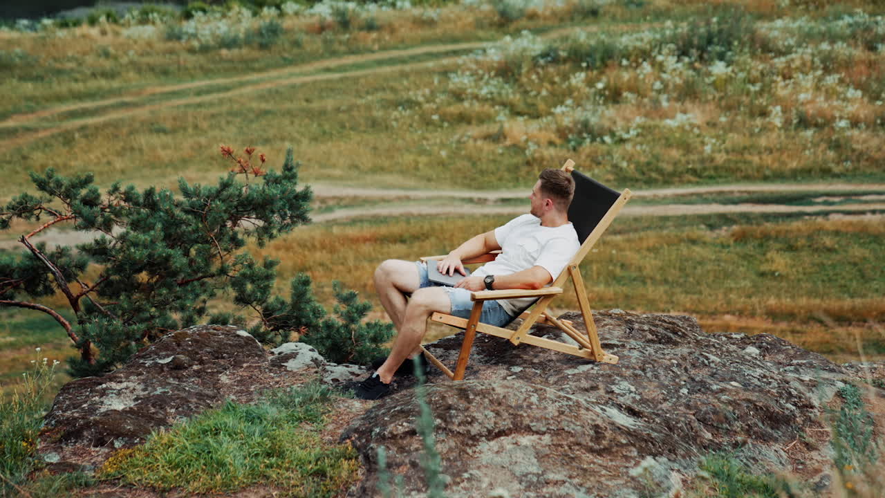 Pensive relaxed male sits in a folding chair on the rock. Man looks around enjoying the view after work on laptop. High angle view.