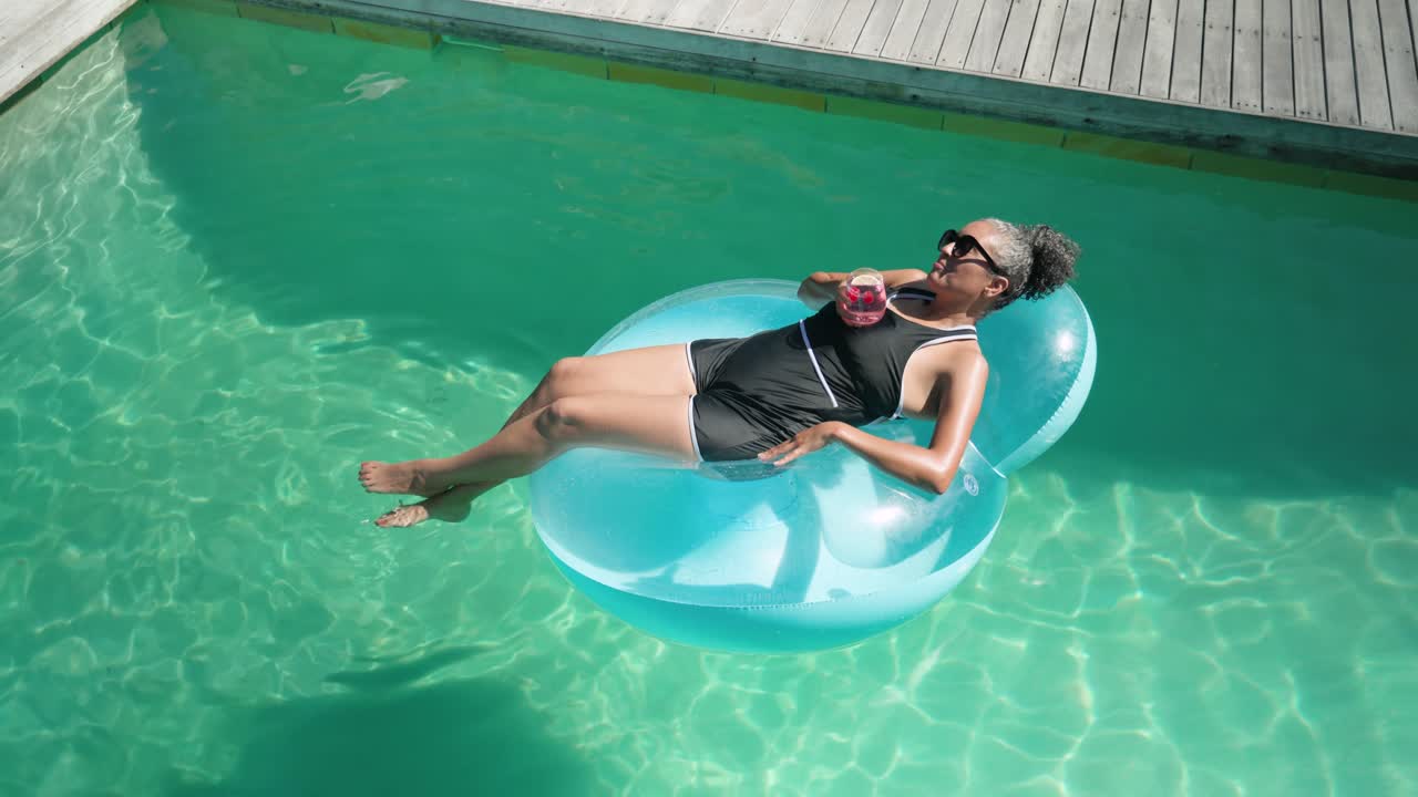 African American woman sunbathing on float in pool sipping pink drink balancing after raft drift