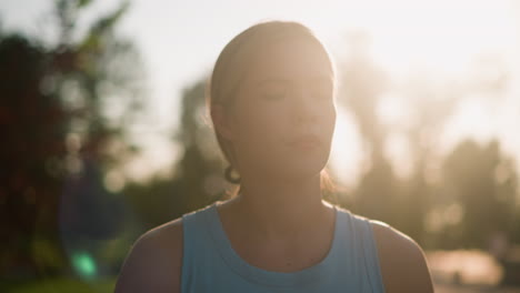 close-up of young woman looking pensive with sunlight glowing around her, creating warm, radiant backlit effect, her hair tied back, blurred natural background of trees