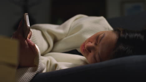 close up of woman spending evening at home lying on sofa with mobile phone scrolling through internet or social media 10