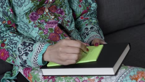 medium close up of young asian indian female's hand in salwar kameez writing down notes with pen sitting down indoors