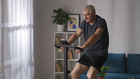 Mann-Mittleren-Alters-Tritt-Zu-Hause-In-Die-Pedale-Eines-Modernen-Heimtrainers-Und-Sieht-Sich-Auf-Dem-Bildschirm-Ein-Video-An,-Um-Cardio-Training-Zu-Absolvieren-Und-Dabei-Körperlich-Aktiv-Zu-Bleiben