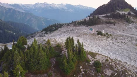 Tiro-De-Dron-Aéreo-De-Inclinación-Lenta-De-Pinos-Y-Cabaña-Solitaria-Al-Atardecer-En-El-Monte-Brew-Con-Montañas-Nevadas-En-Canadá-Bc-4k