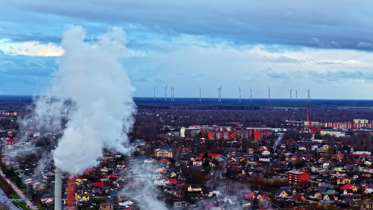 Un'alta ciminiera rilascia un denso pennacchio di fumo sopra una città, con file di turbine eoliche visibili all'orizzonte in lontananza - Campo lungo