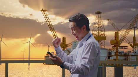 side view of an asian male professional worker standing with his smartphone with offshore hydrogen production, high-tech industrial facility, he raises his fist up with screaming goal after check on the smartphone