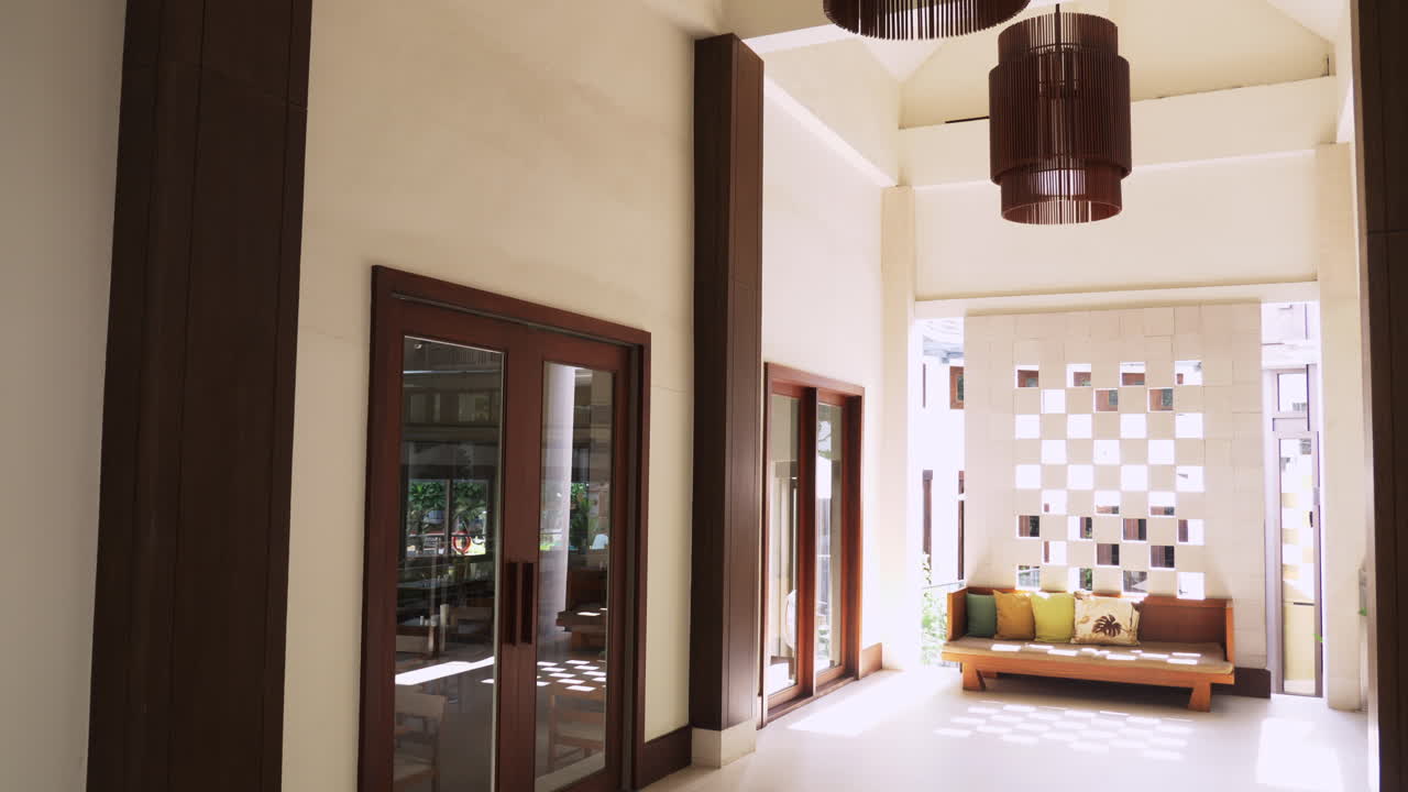 Soffitto e lampadari si rivelano mentre la telecamera si inclina verso il basso, mostrando una vista più ampia di una hall, finestre, porte e un divano con cuscini decorativi.