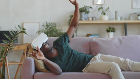 Aufgeregter-Afroamerikanischer-Mann,-Der-Auf-Dem-Sofa-Liegt-Und-Ein-VR-Headset-Benutzt