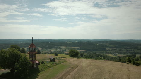 Toma-Panorámica-De-Una-Colina-Con-Una-Pequeña-Torre-En-La-Parte-Superior-Y-Un-Bosque-Con-árboles-Y-Campos-Agrícolas-De-Hierba-Verde-A-Su-Alrededor-Cielo-Azul-Y-Colinas-En-El-Fondo-En-Eslovenia,-Toma-Aérea-De-Drones