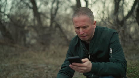 a close view of a man wearing a green jacket and blue jeans sitting in a forested area, deeply focused on his smartphone, with a gun in his hand facing down