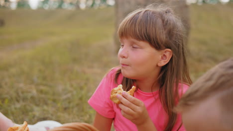 little kid in pink top enjoys snacks outdoors in open field while partial head view of another boy eating pastries nearby, capturing joyful moments in a peaceful rural landscape