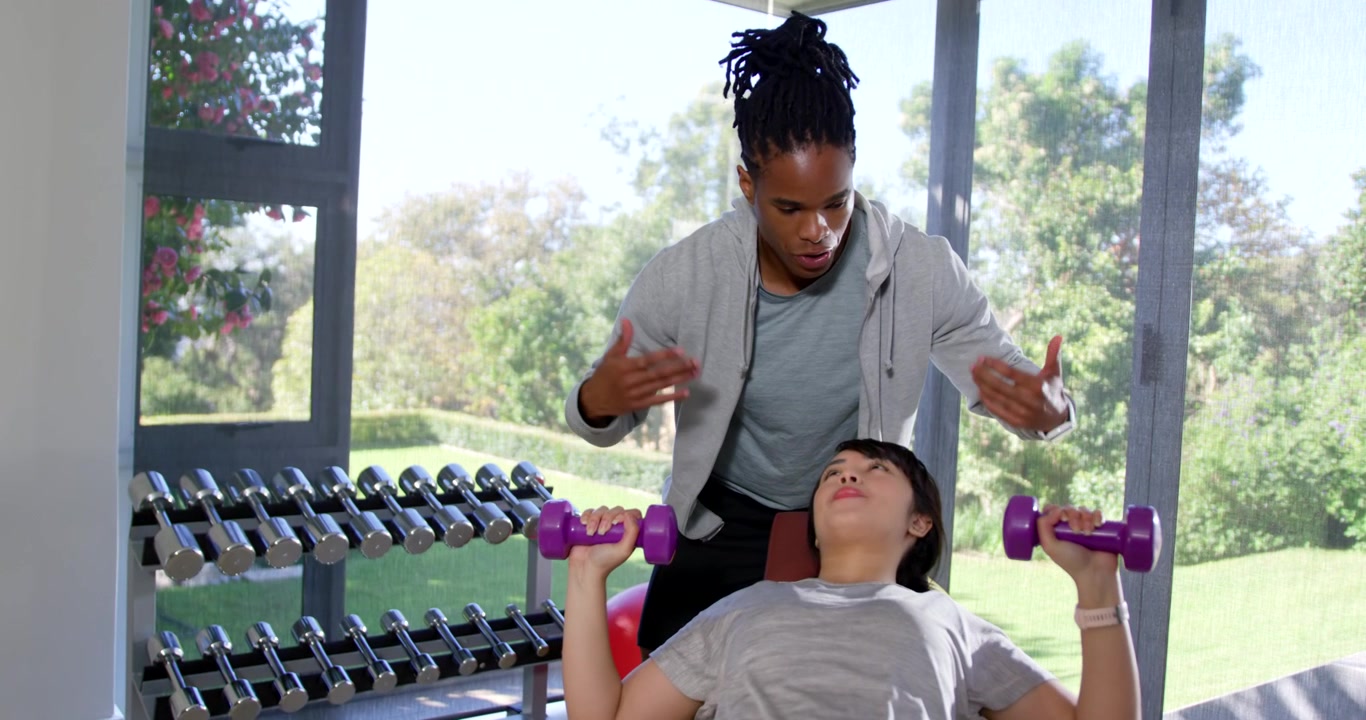 African American trainer cueing guiding hands for form as Asian trainee pressing dumbbells on bench