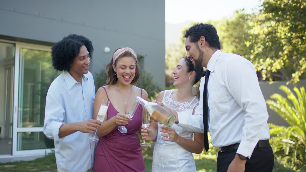 Diverse friends bride in white smiling lawn man right tilting bubbly bottle pouring flutes toasting