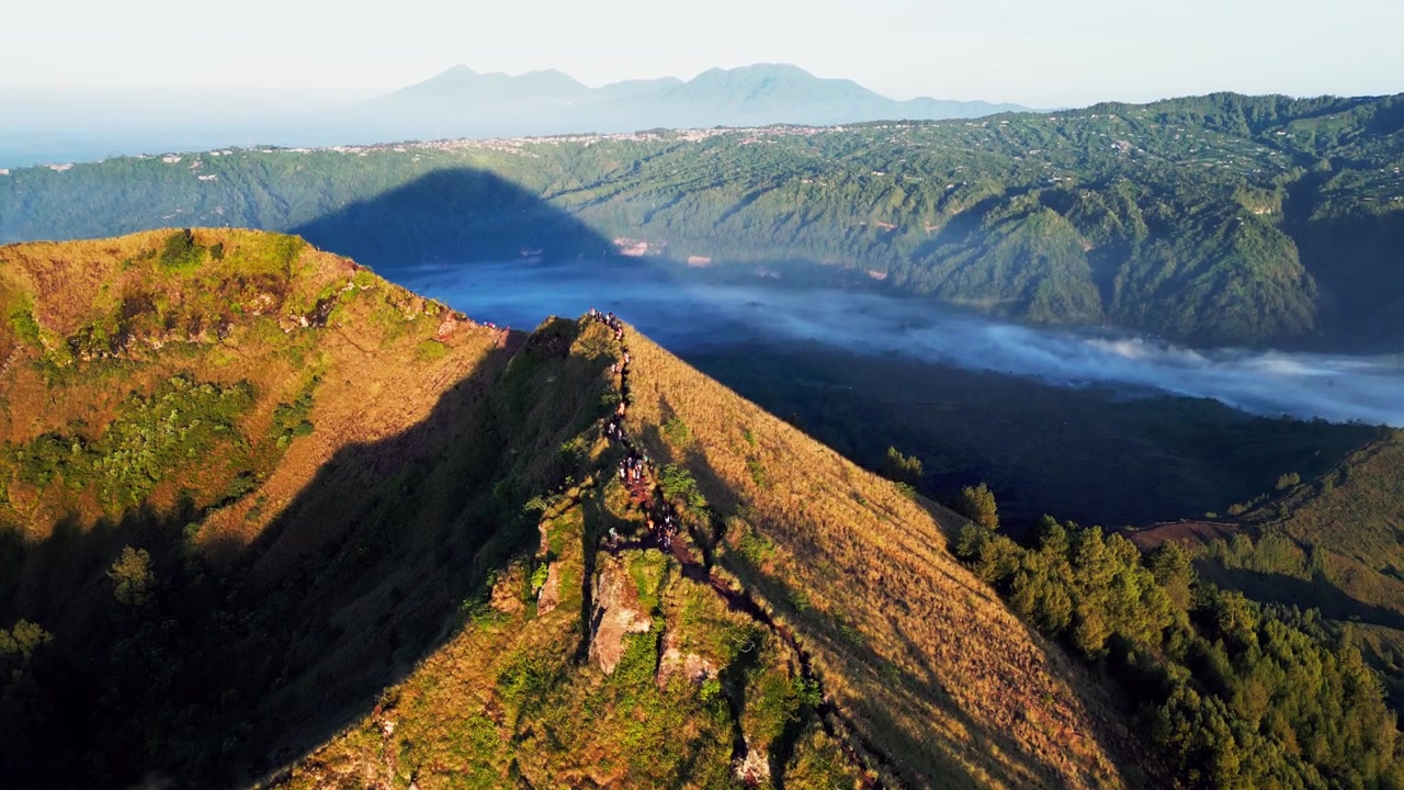 Sentiero di cresta montano con escursionisti che camminano lungo un percorso esposto, luce dell'alba che modella il terreno accidentato e viste della caldera sopra la foschia mattutina a Bali, Indonesia, riprese aeree con drone