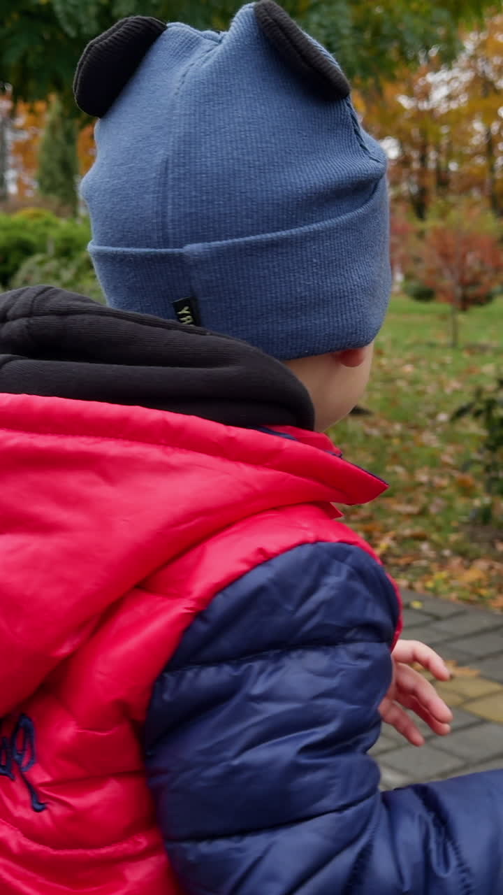 Lovely peaceful Caucasian toddler in cap and red jacket is in the park. Baby boy walking in autumn park. Vertical video