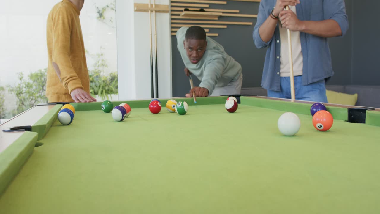 Encouraged by friend gesture, African American man taking shot at rec room pool table striking ball