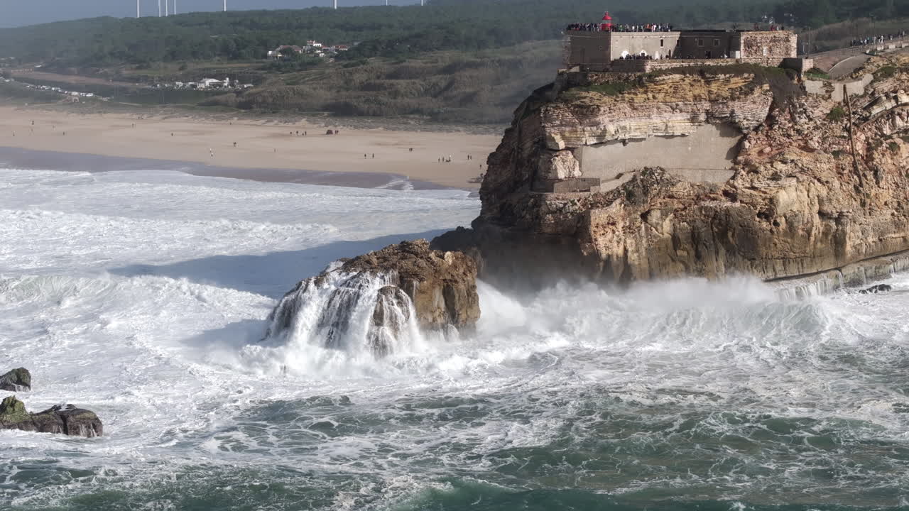 Beautiful aerial drone shot of big waves breaking on rocks and cliffs on a day with giant waves in Nazaré, Portugal, Europe. Farol da Nazare lighthouse and wind turbines visible