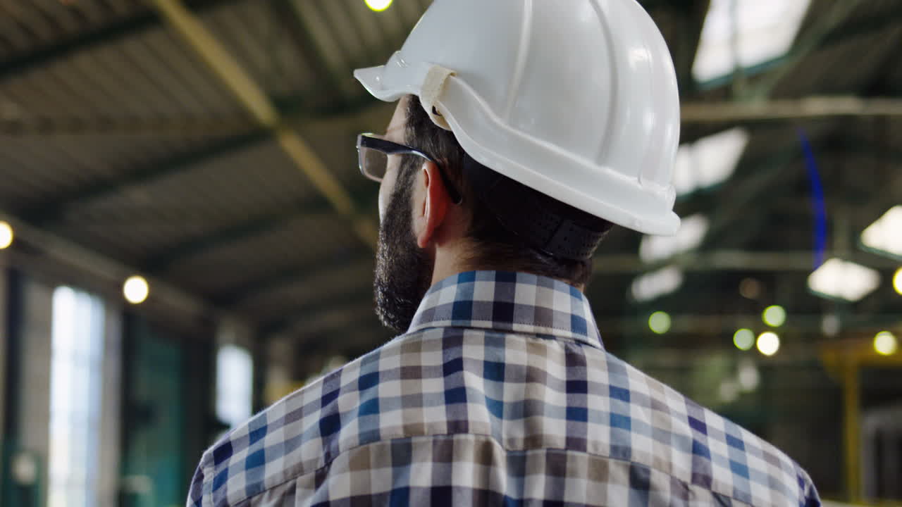 Rear view of male worker in a plaid shirt and a helmet walking in a factory