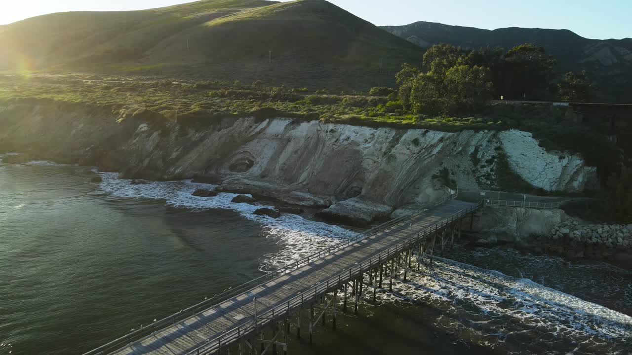 el sol se levanta sobre las montañas proyectando luz sobre el muelle público y los viejos caballetes del ferrocarril sobre la playa con colinas verdes cubiertas de hierba detrás, órbita aérea de gaviota beach, california