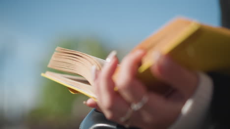 primer plano de la mano con uñas bien pulidas adornadas con anillos volviendo a través de las páginas del libro de portada amarilla contra un suave fondo borroso, mostrando un momento de lectura suave y reflexivo