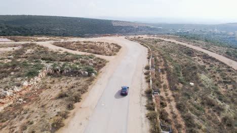 Luftdrohne-Schoss-Tagsüber-In-Katzir,-Israel,-Nach-Einem-Schwarzen-Auto-Durch-Einen-Fichtenwald-Auf-Hügeligem-Gelände