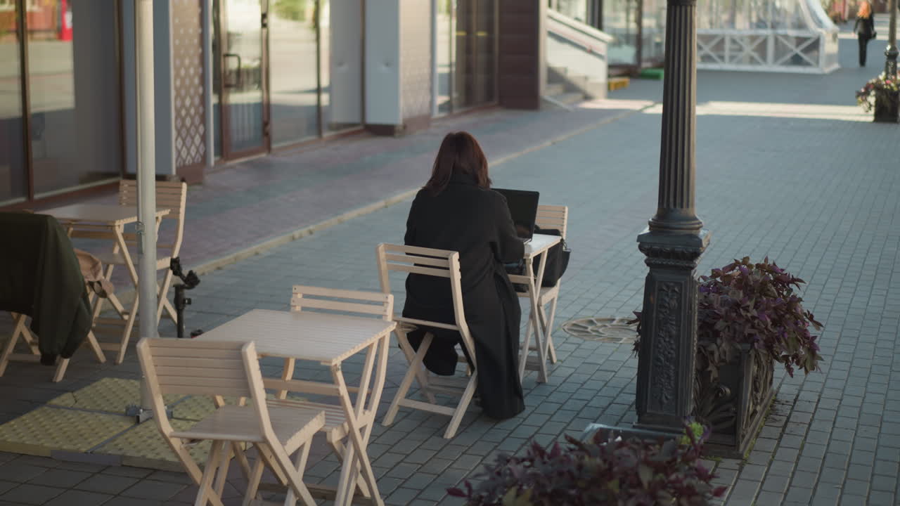 vista trasera de una mujer escribiendo en una computadora portátil al aire libre, vista parcial de alguien con un traje negro caminando en la distancia, con plantas y sillas de madera alrededor