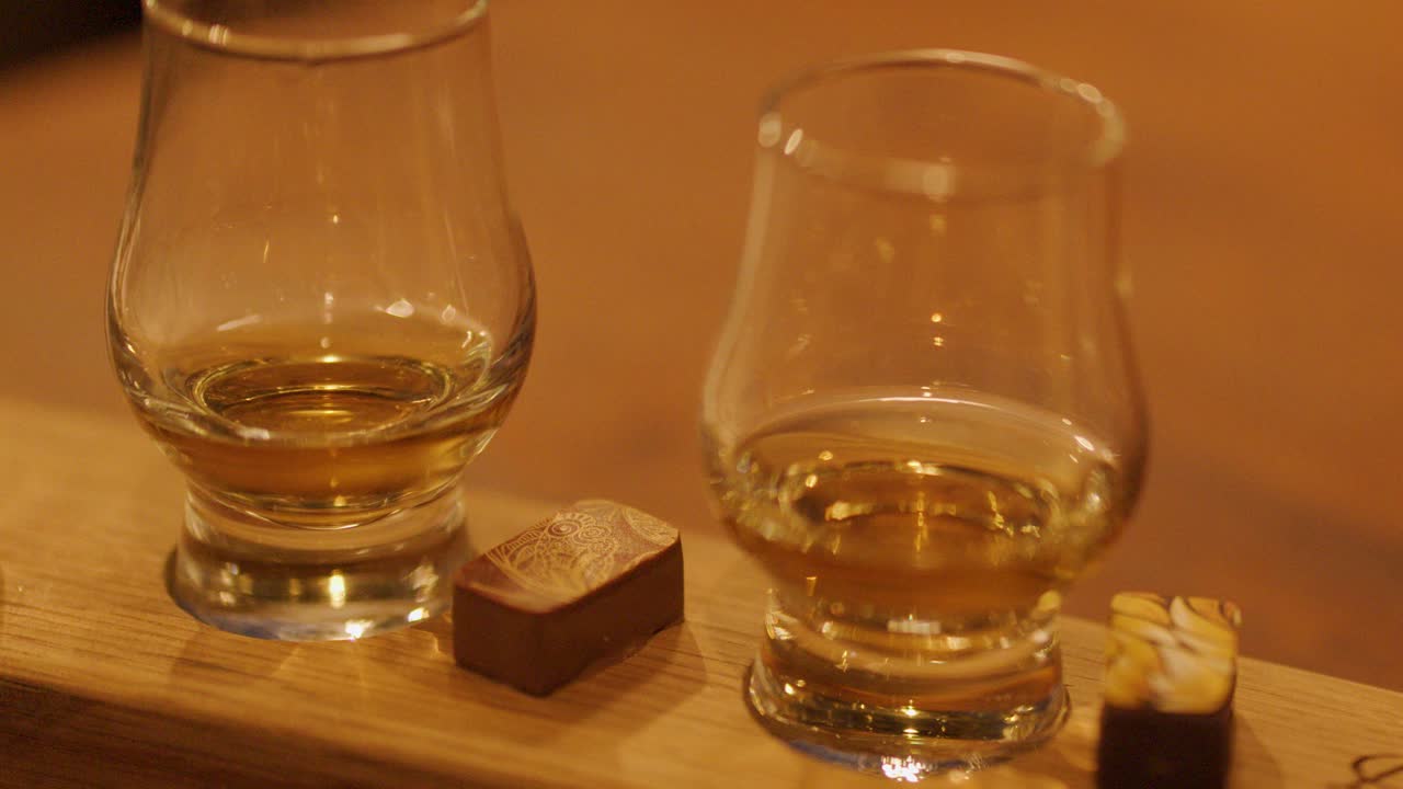A hand sets a whiskey glass next to chocolate pieces on a wooden tray in warm, ambient lighting. Close-up, steady camera, inviting mood