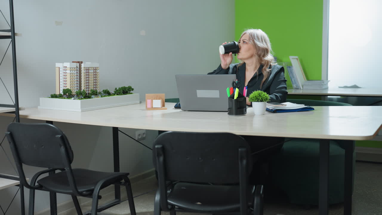 Real estate mogul seated at desk working on laptop pauses to sip coffee and resumes typing, surrounded by modern office setting, architectural model