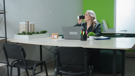 real estate mogul seated at desk working on laptop pauses to sip coffee and resumes typing, surrounded by modern office setting, architectural model