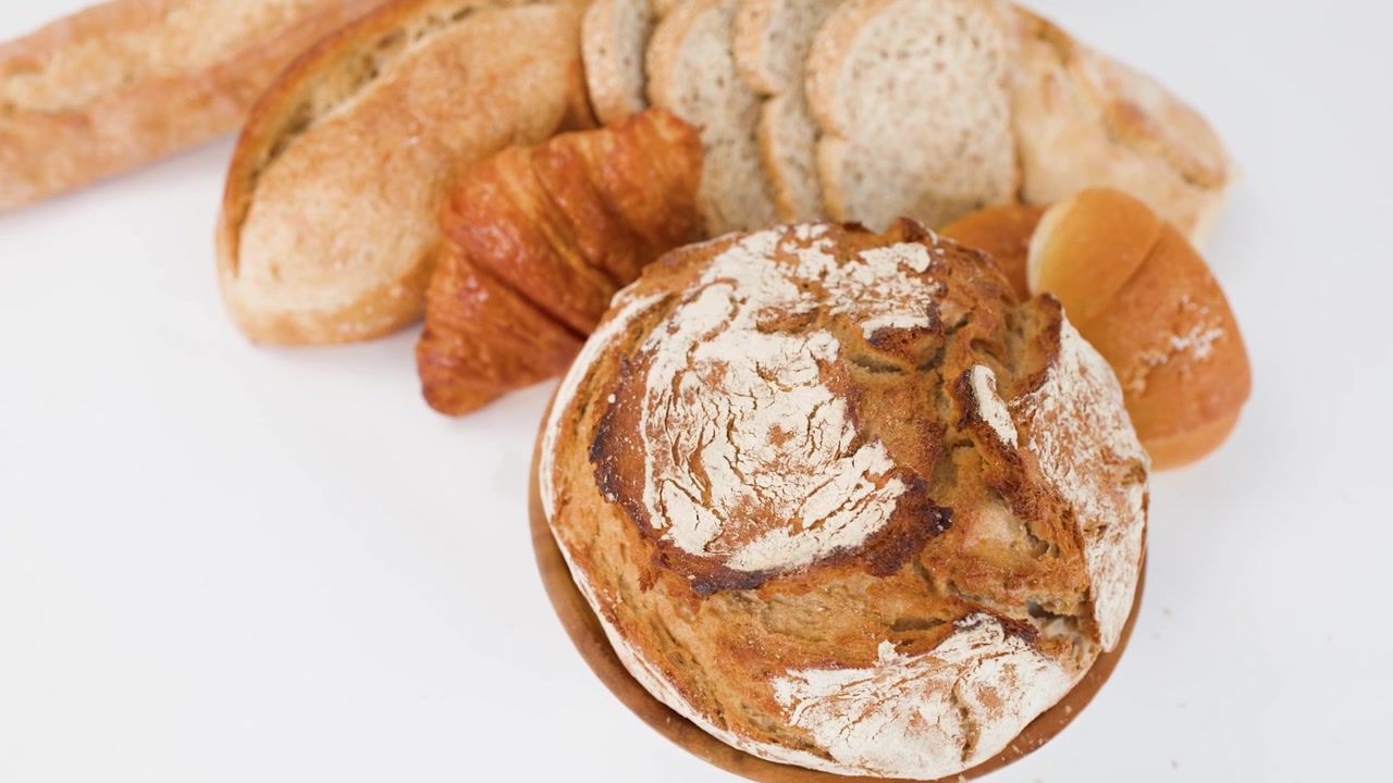 Lento zoom indietro rivela una pagnotta di pane a lievitazione naturale, un croissant e fette di baguette sotto una luminosa illuminazione da studio