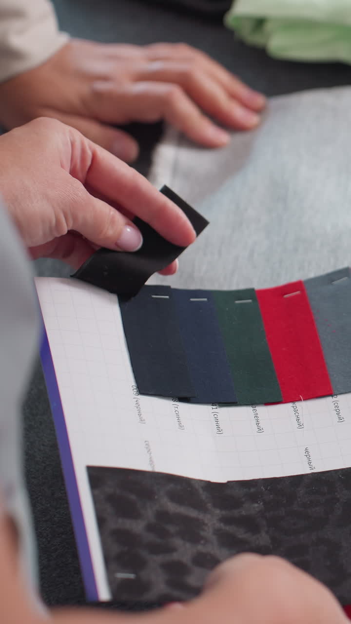 Manos comparando muestras de tela oscura en una tabla, consultor textil evaluando el tono y la textura con pruebas precisas con el pulgar, muestras de tapicería y prendas de vestir en capas, luz de estudio brillante, concentración