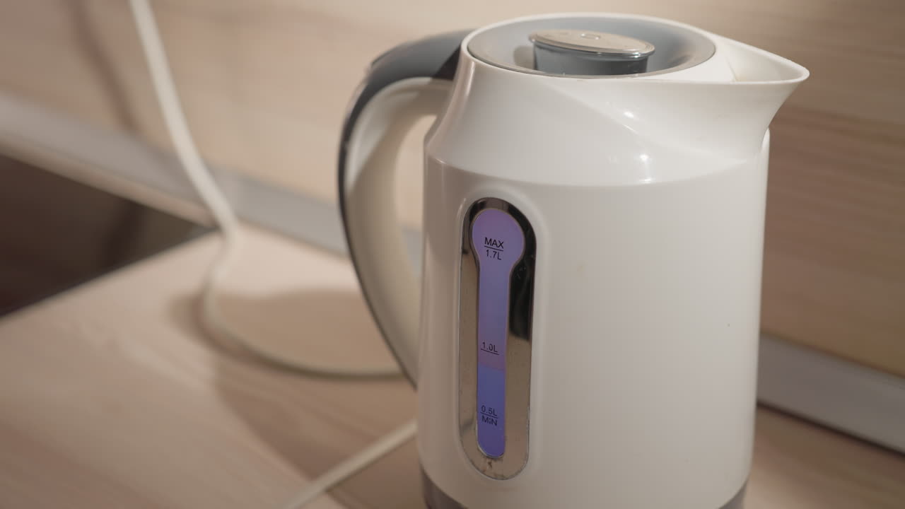 Close-up shot of electric kettle with visible water level indicator in modern minimalist kitchen. Soft lighting highlights sleek design and clean lines of kettle and kitchen countertop