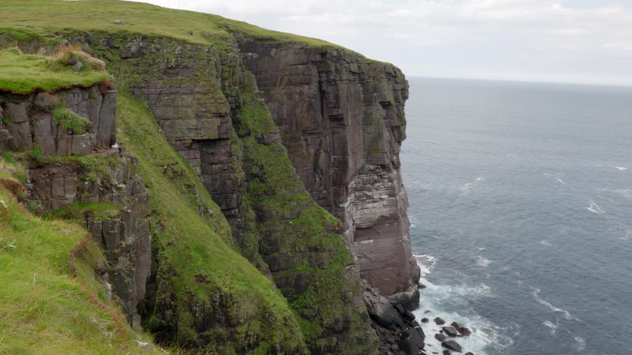 A slow tilting and panning shot, rising from a rocky bay as waves lap against a tall sea cliff in the ocean while seabirds fly around grassy cliffs in a seabird colony