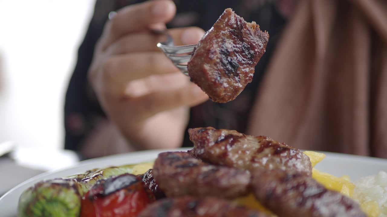 A close up of a plate of Turkish koftes, with grilled vegetables, rice and potatoes.  A person is holding a fork with a kofte on it.