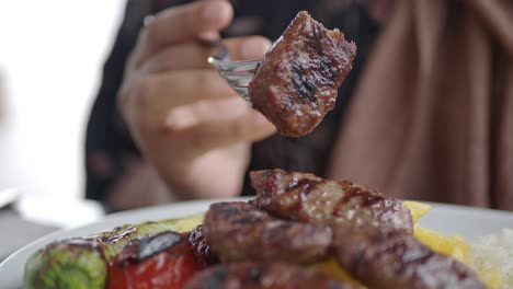 a close up of a plate of turkish koftes, with grilled vegetables, rice and potatoes.  a person is holding a fork with a kofte on it.